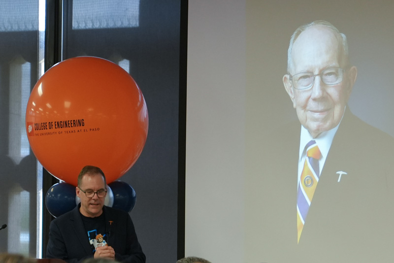 Attendee wearing a name badge stands beside a banquet table during a UTEP Engineering homecoming gathering while other attendees applaud, with orange and blue table decorations, coffee cups, and event materials visible throughout the room.